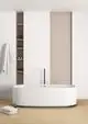 Freistehende Badewanne von Ideal Standard in schlichtem Weiß, mit schwarzem Wasserhahn hinter der Wanne, vor hellen Wänden und einem Regal mit Pflegeprodukten.